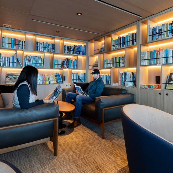 Two people sit in an on ship library reading, with a backdrop of books on bookshelves