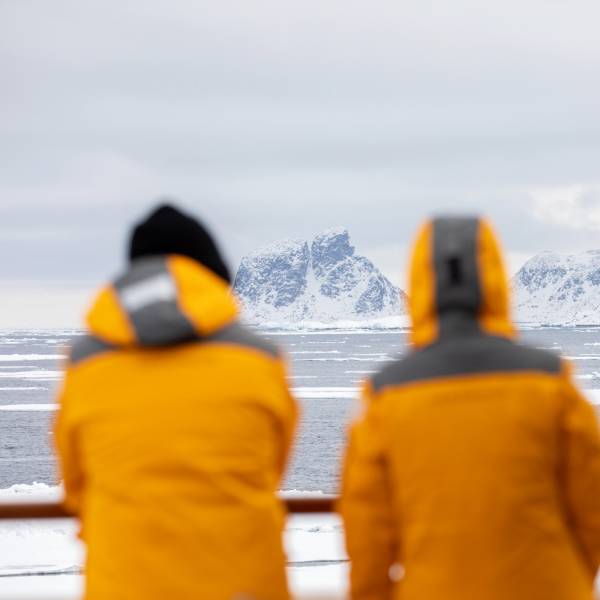 Two people stand on a ship with their backs to the camera, wearing their Quark Expeditions parkas, looking out onto an icy landscape