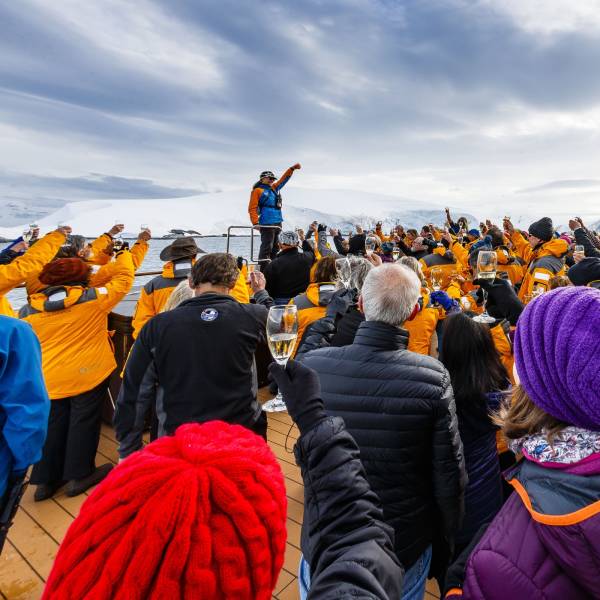 Guests on the deck of a polar expedition ship, wearing Quark Expedition parkas, gathered around an expedition leader while everyone raises a glass of champagne in celebration against a backdrop of ice