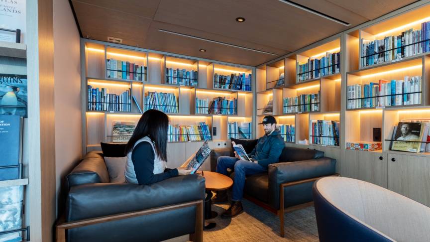 Two people sit in an on ship library reading, with a backdrop of books on bookshelves