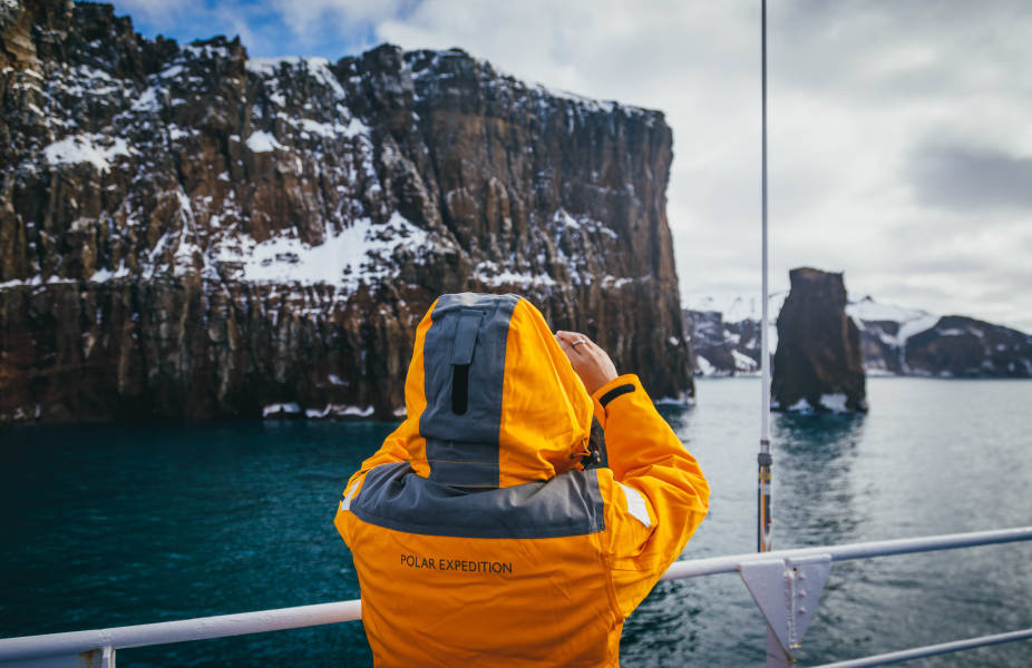 Guest in yellow parka holds their smartphone in front of their face as they take a photo of the sights off the ship.