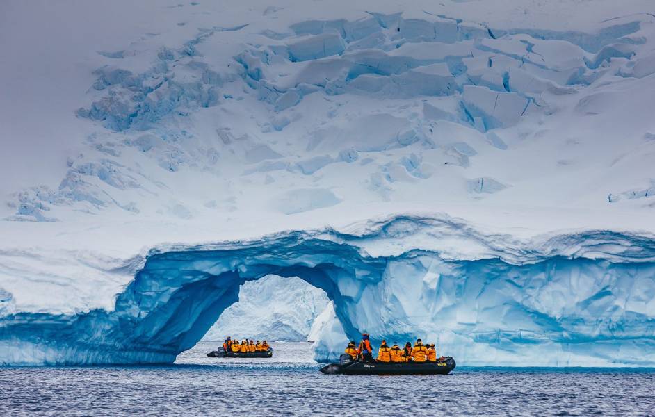 A zodiac boat is spotted through the arch of an iceberg near Cuverville Island. The zodiac boat appears to be close to the iceberg as a result of telephoto lens compression.