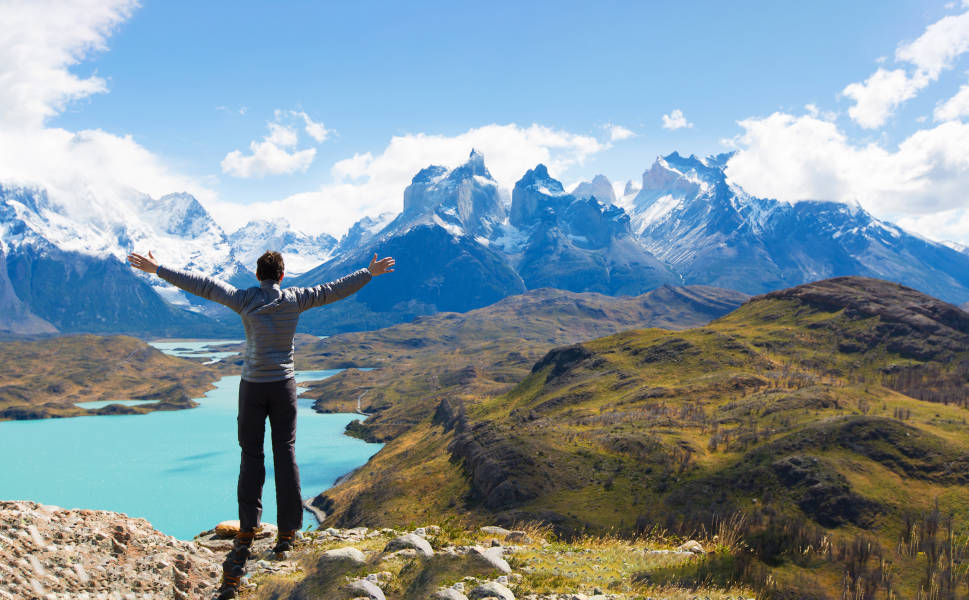 A guest stands on a cliff's edge with arms extended in front of clear light blue waters and lush green grass, with the  mountain peaks of Cuernos del Paine in the background. The photo was taken at Torres del Paine National Park, Chile.