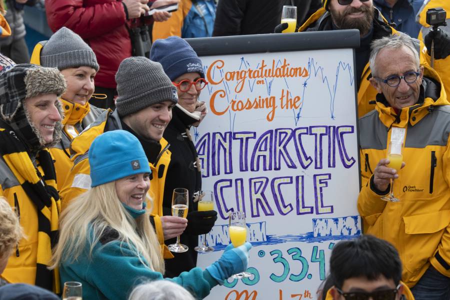 Guests celebrate crossing the Antarctic Circle with sparkling beverages and taking photos on the deck of the ship.