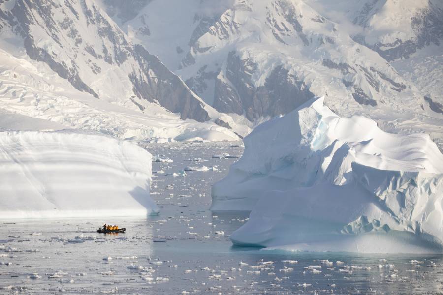 A zodiac boat is spotted cruising near grounded icebergs. The zodiac boat appears to be close to the iceberg as a result of telephoto lens compression.