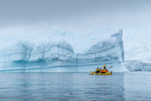 Two guests kayaking through frigid waters in Antarctica. A big iceberg can be seen in the background