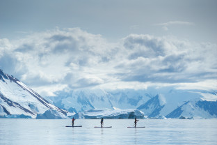 Guests go standup paddleboarding n the Antarctica Peninsula. Snow covered mountains are seen in the distance.