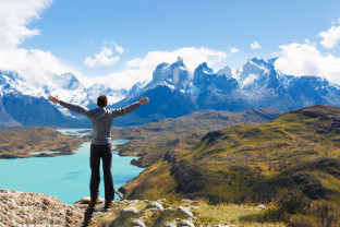 A guest stands on a cliff's edge with arms extended in front of clear light blue waters and lush green grass, with the  mountain peaks of Cuernos del Paine in the background. The photo was taken at Torres del Paine National Park, Chile.