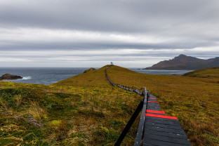 The image shows a long boardwalk stairway with the Cape Horn Monument at the end. The monument pays homage to sailors that perished attempting to round Cape Horn and depicts an albatross in flight.