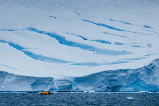 Zodiac boat in front of iceberg. Guests in yellow parkas can be seen taking photos while their Expedition Guide, in red, drives the boat.
