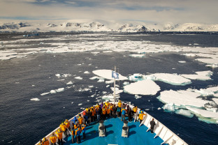 Guests in yellow parkas stand at the bow of their vessel as it cruises through sea ice in the Antarctic Peninsula.