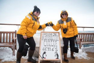 Two guests in yellow parkas pose for a photo next to a board that includes the coordinates of their ship, celebrating officially crossing the Antarctic Circle.