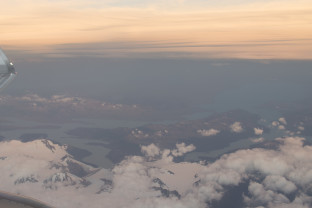 View from the window of an aircraft featuring its wing and a small cloud cover over the partially ice-covered peaks near Punta Arenas, Chile.